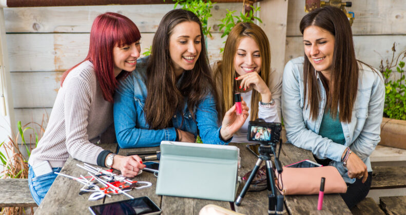 This image has four women sitting around a table that has a tablet, camera and other photo props.